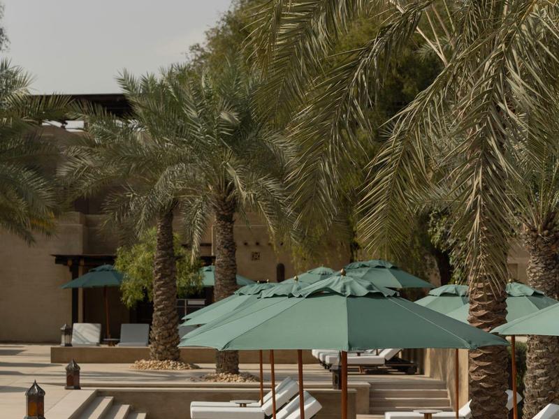 White lounge chairs under green umbrellas next to palm trees on a hotel terrace.