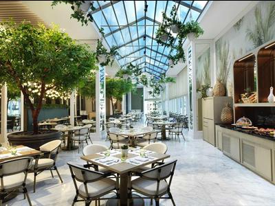 Heller Wintergarten mit großen Bäumen, runden Tischen und Stühlen, Glasdach und Buffetbereich.