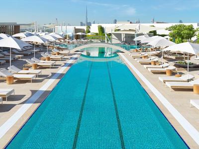 Moderner Außenpool mit blauen Fliesen, weißen Liegen und Sonnenschirmen in sonniger Stadtumgebung.