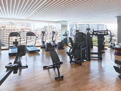 Jasna siłownia z drewnianą podłogą i widokiem na miasto przez duże okna.