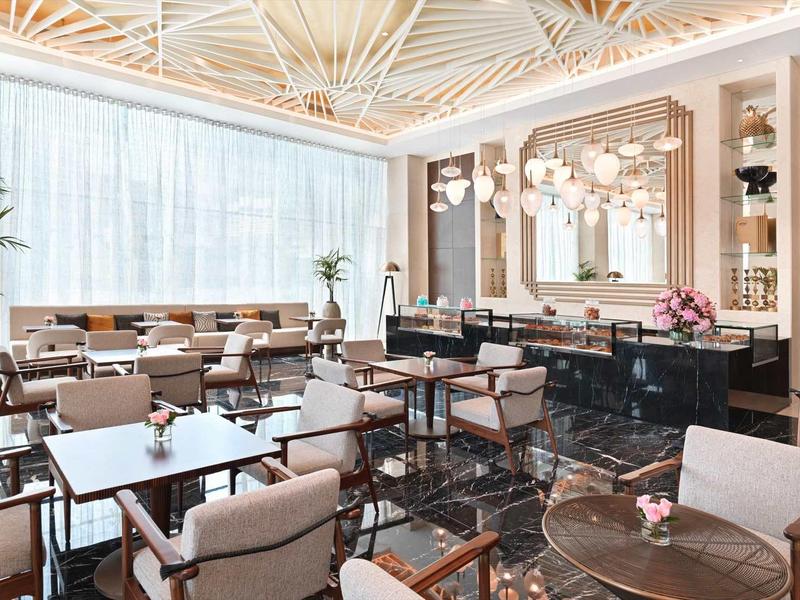 Moderner, heller Speiseraum mit weißen Vorhängen, Holztischen und beigen Sesseln. Deckendekor auffällig.