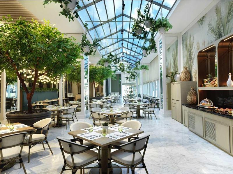 Heller Wintergarten mit großen Bäumen, runden Tischen und Stühlen, Glasdach und Buffetbereich.