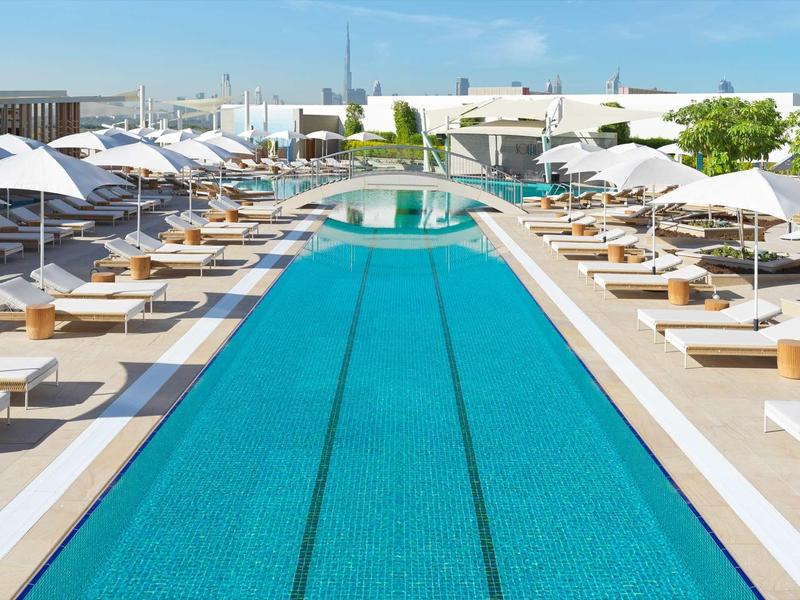 Moderner Außenpool mit blauen Fliesen, weißen Liegen und Sonnenschirmen in sonniger Stadtumgebung.