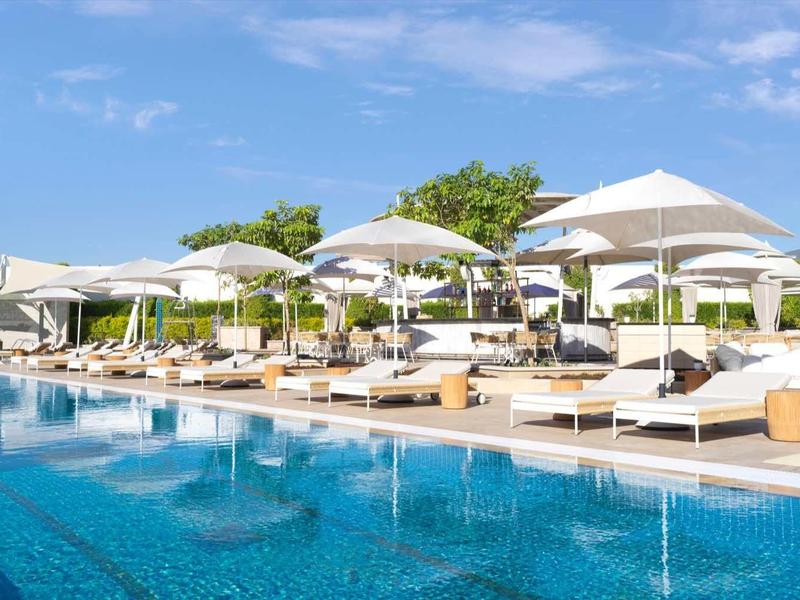 Großer Außenpool mit blauer Wasseroberfläche, weißen Liegen und Sonnenschirmen unter klarem Himmel.