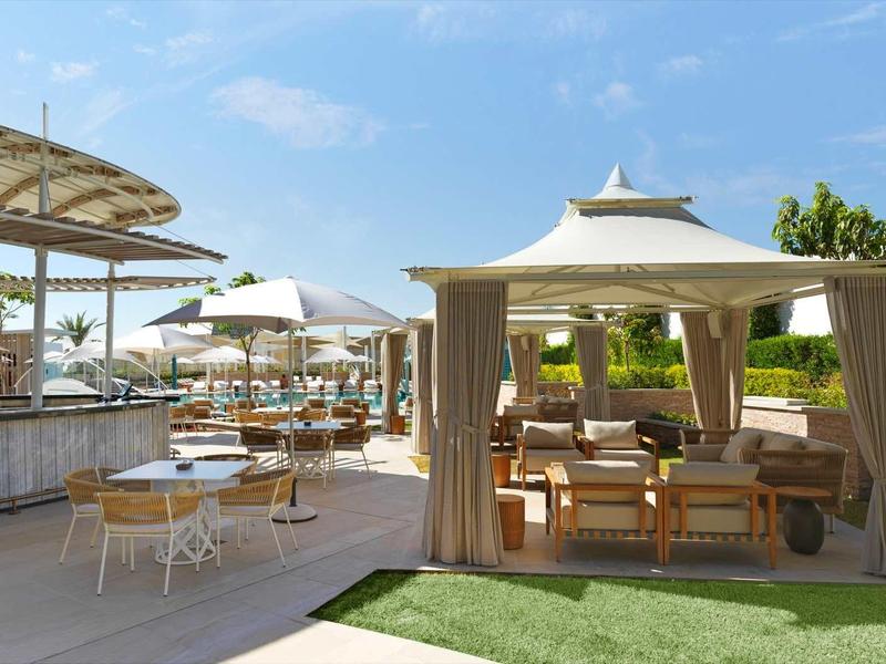 Outdoor-Café mit Tischen, Stühlen, Sonnenschirmen und überdachtem Sitzbereich bei blauem Himmel.