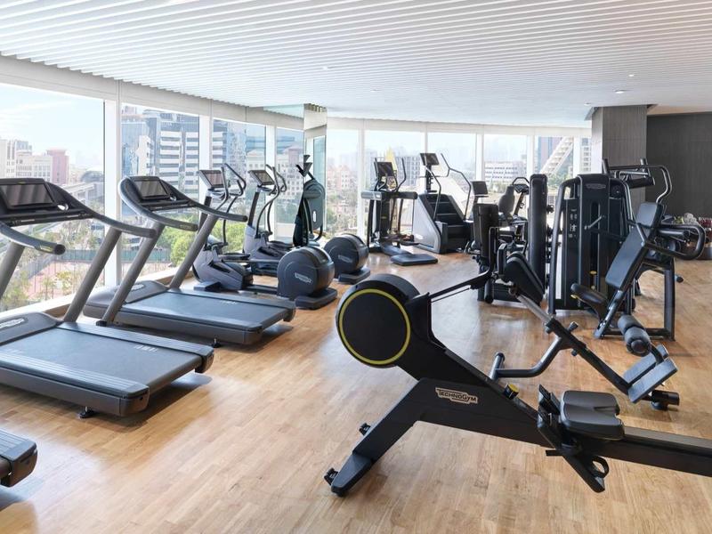 Heller Fitnessraum mit Holzfußboden, Laufbändern, Rudergerät und Kraftstationen vor großen Fenstern.