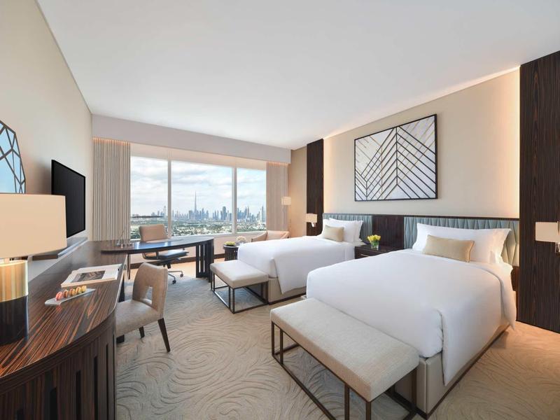 Hell eingerichtetes Hotelzimmer mit zwei Betten, Schreibtisch, Fernseher und großem Fenster mit Stadtblick.