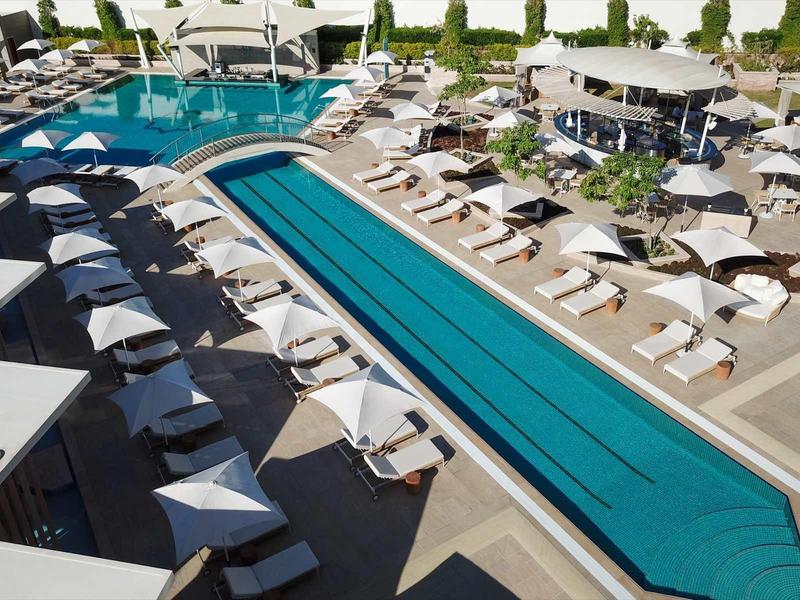 Großer moderner Außenpool mit Liegestühlen, Sonnenschirmen und umliegender Terrasse bei Tageslicht.