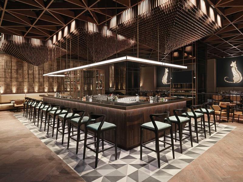 Moderner Barbereich mit dunklem Holz, hohen Stühlen und geometrischem Bodenmuster in warmem Licht.