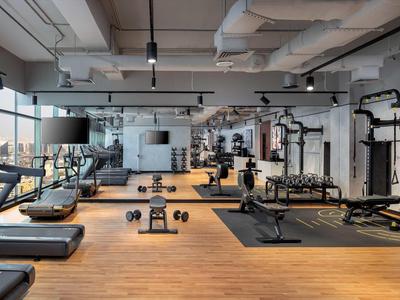 Moderner Fitnessraum mit Geräten, Hanteln, Holzfußboden und großen Fenstern für Tageslicht.