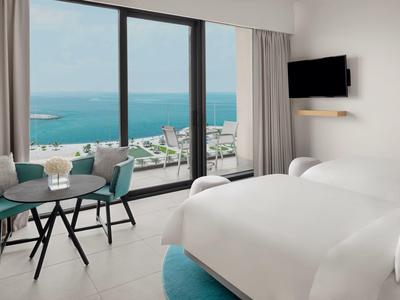 Helles Hotelzimmer mit großem Fenster, Meerblick, zwei Betten, Tisch, Stühlen und Wand-TV.