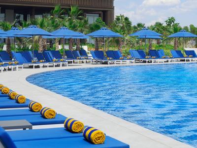 Großer Pool mit blauen Liegestühlen und Sonnenschirmen, umgeben von Palmen und klarem Himmel.