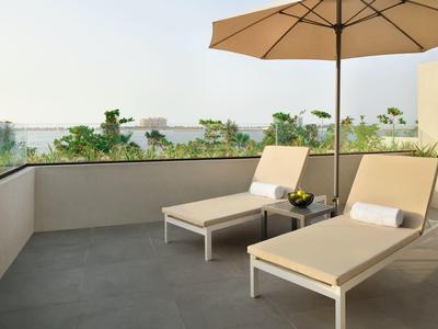 Terrasse mit zwei beigen Liegestühlen, Sonnenschirm und Blick auf grüne Bäume im Hintergrund.