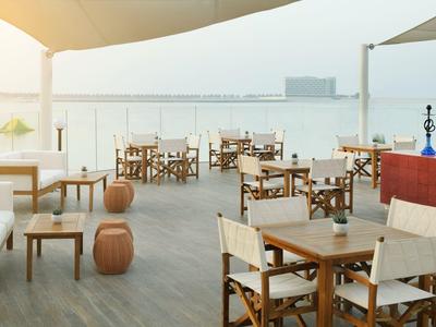 Leerer Außenbereich eines Cafés mit Holzboden, Stühlen, Tischen und Blick auf Wasser bei Sonnenuntergang.