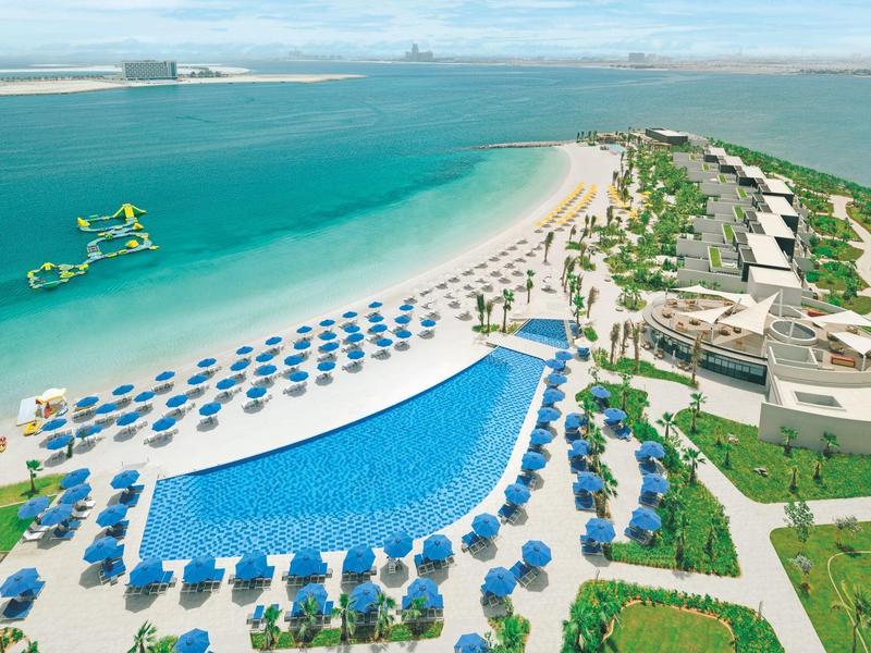 Großer Pool mit blauen Sonnenschirmen neben Strand und Meer in einem Resort.