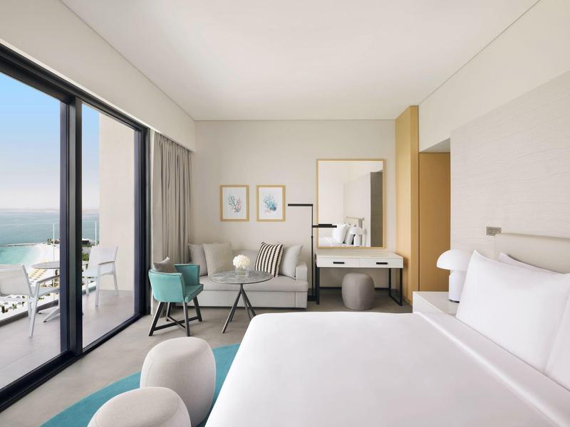 Helles Hotelzimmer mit großem Bett, Sitzbereich und Meerblick durch bodentiefe Fenster.