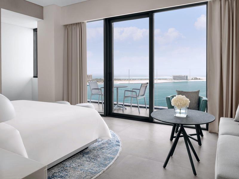 Modernes helles Schlafzimmer mit Balkon, Meerblick, Sofa, Bett und rundem Tisch.