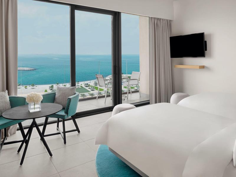 Helles Hotelzimmer mit großem Fenster, Meerblick, zwei Betten, Tisch, Stühlen und Wand-TV.