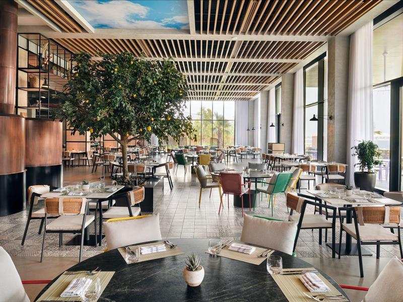 Heller, moderner Gastraum mit großen Fenstern, Pflanzen und verschiedenen Tischen und Stühlen.