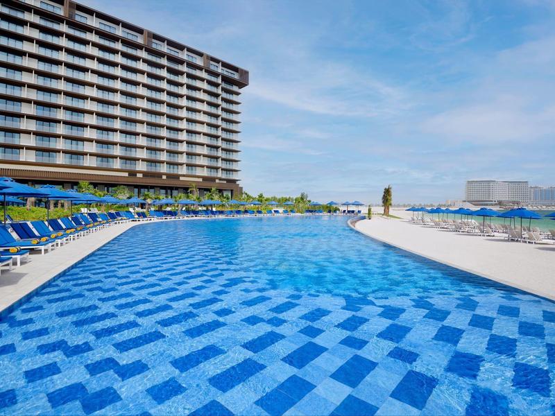 Moderner Außenpool mit blau karierten Fliesen, Liegestühlen und großen Gebäuden im Hintergrund.