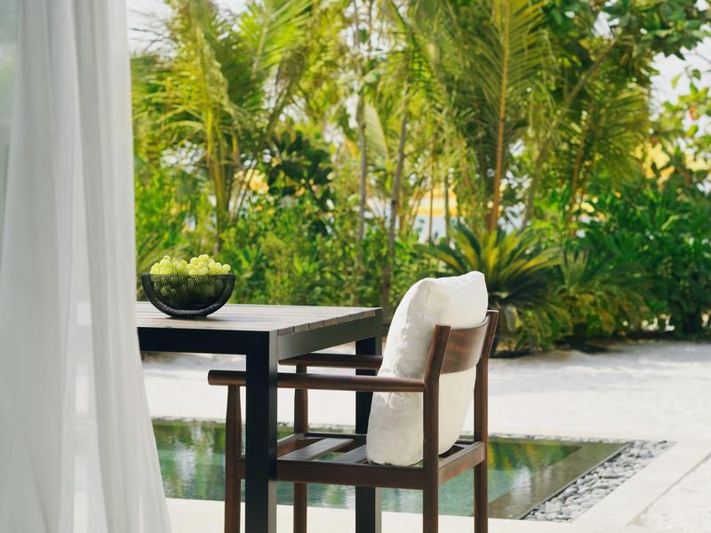 Moderner Stuhl mit Polster und Holzrahmen neben schwarzem Tisch und Pflanzen im hellen Außenbereich.