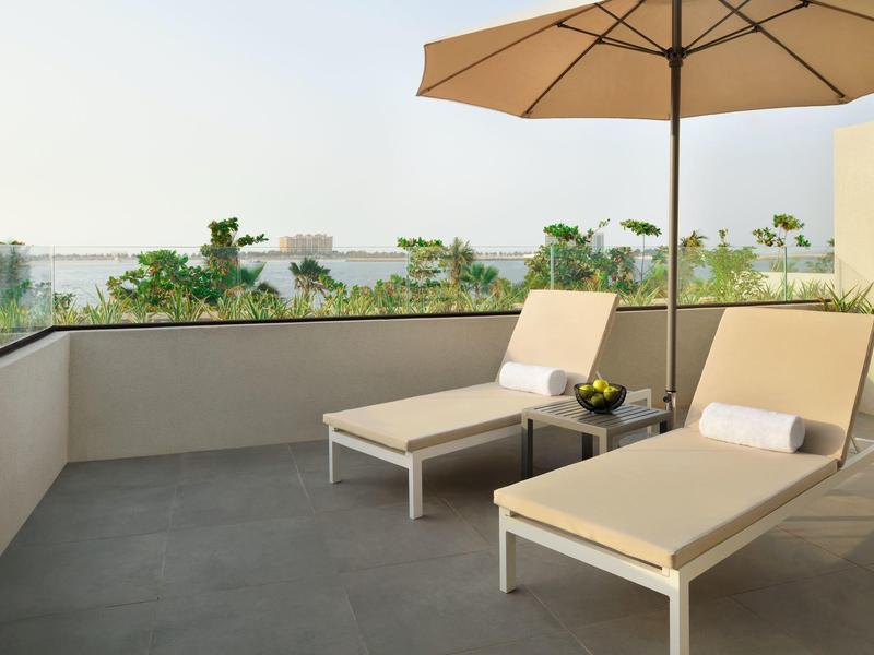 Terrasse mit zwei beigen Liegestühlen, Sonnenschirm und Blick auf grüne Bäume im Hintergrund.