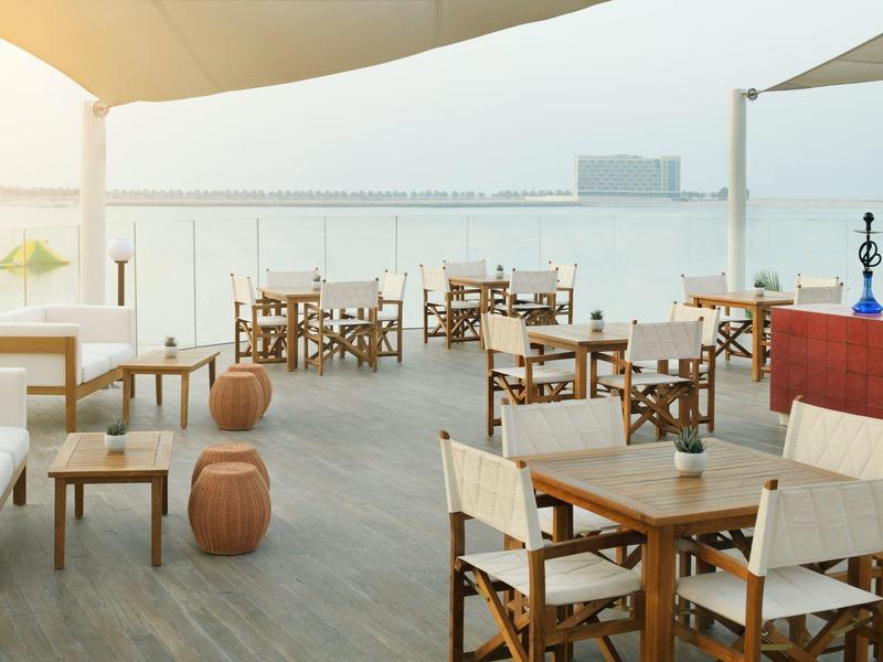 Leerer Außenbereich eines Cafés mit Holzboden, Stühlen, Tischen und Blick auf Wasser bei Sonnenuntergang.