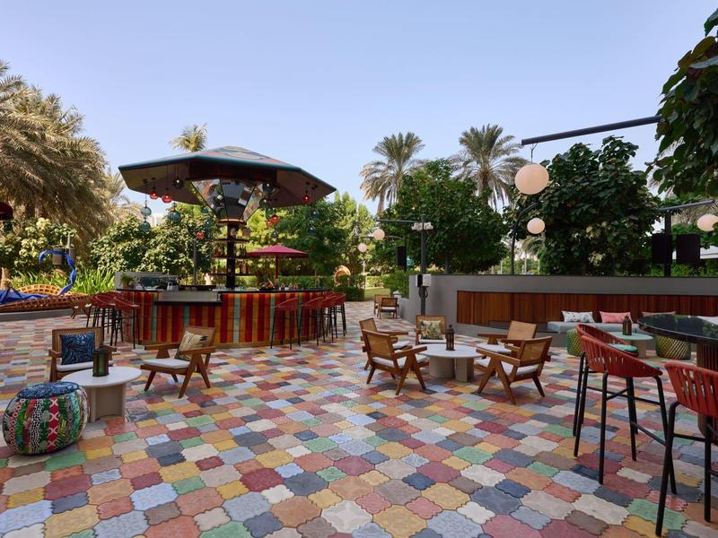 Colorful outdoor bar with tiled floor, wooden tables and chairs, umbrellas, and palm trees.