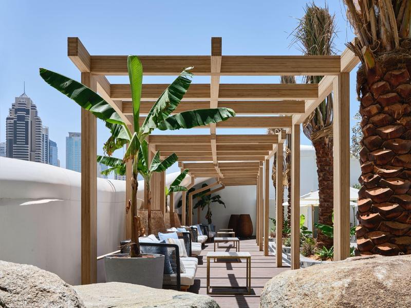 Słoneczny taras z drewnianymi pergolami, roślinami i stołami z krzesłami w miejskim otoczeniu.