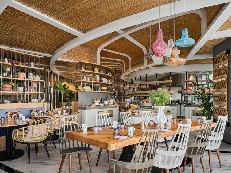 Modern restaurant with wooden tables, white chairs, and colorful pendant lights under a vaulted ceiling.