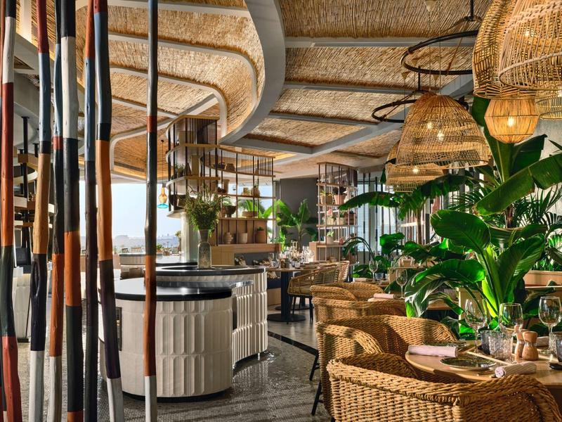 Wnętrze restauracji z okrągłymi stołami, wiklinowymi krzesłami i dużymi roślinami.