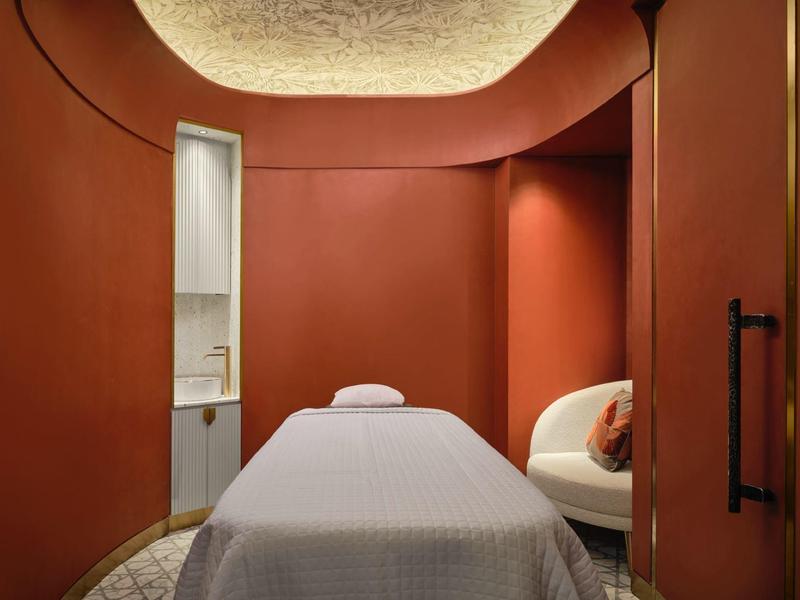 Modern spa treatment room with red walls, massage table, and comfortable chair.