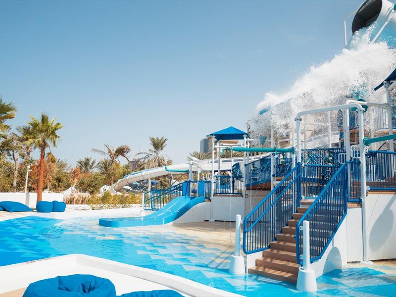 Water park with blue water slides, pool, and palm trees under a blue sky.
