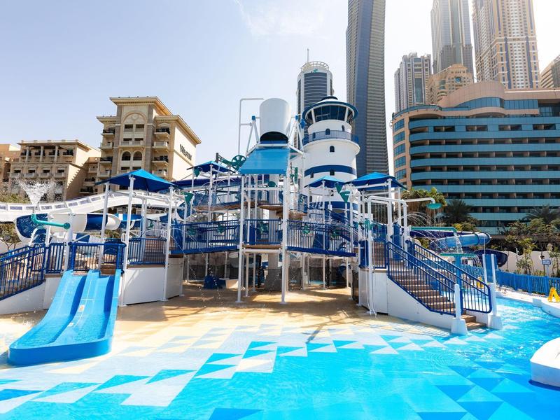 Water park with slides and blue pool in front of modern skyscrapers on a sunny day.