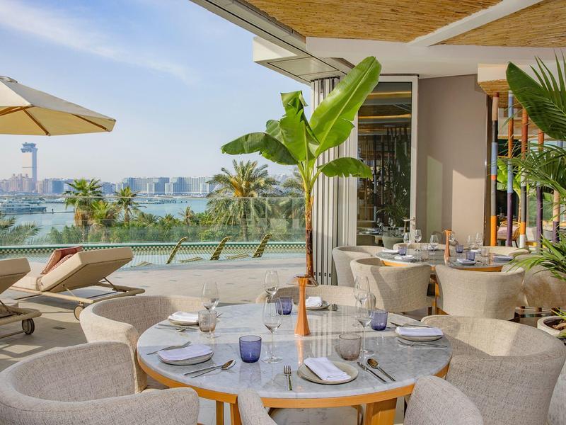 Elegant restaurant with outdoor terrace and sea view, surrounded by tropical plants.