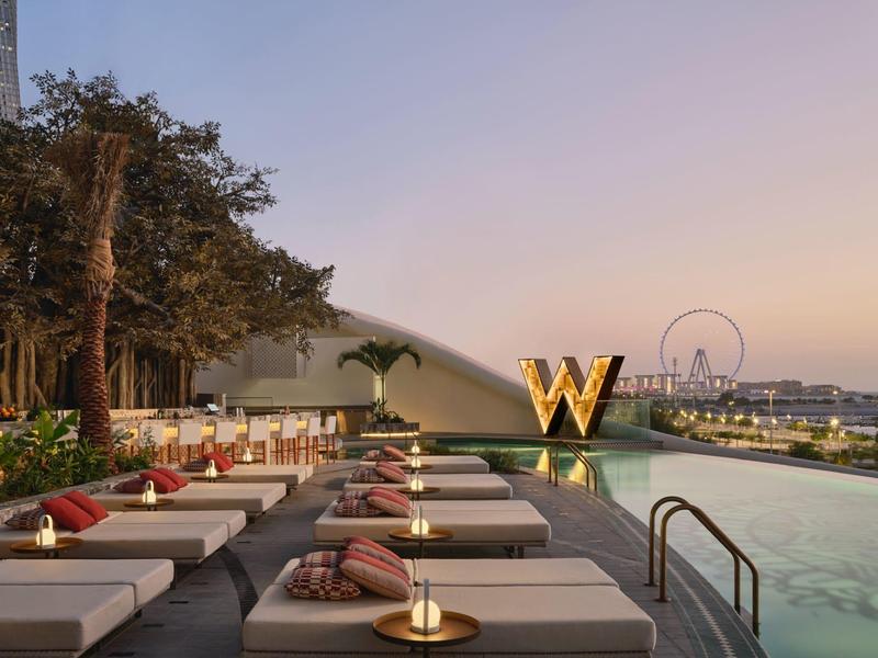 Infinity pool with sunbeds, city and Ferris wheel view at sunset.