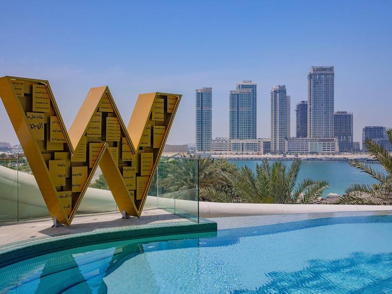 View of a pool with large golden W, skyline and palm trees under blue sky.