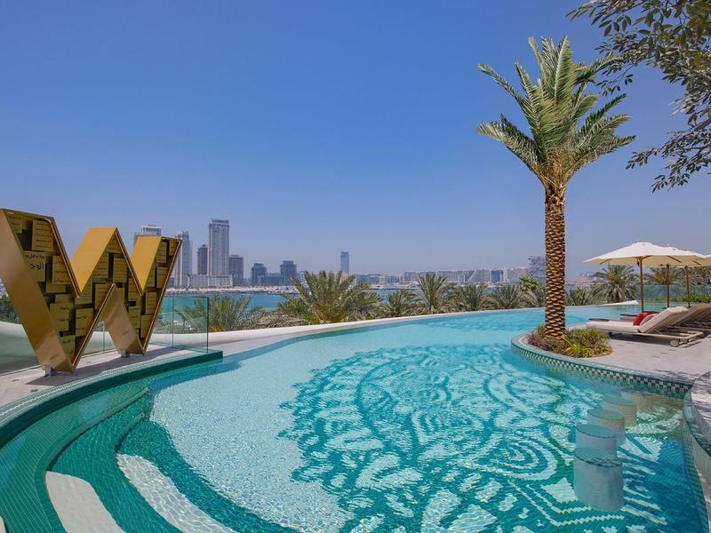 Round outdoor pool with palm trees and city view under sunny weather at luxury hotel.