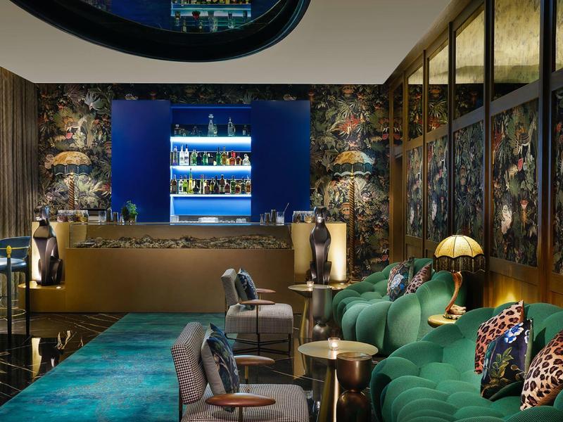 Modern hotel lounge area with green sofas, stylish chairs, and illuminated bar shelves.