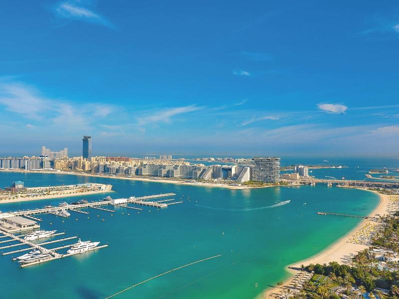 View of a marina with yachts, a beach, and modern buildings under a blue sky.