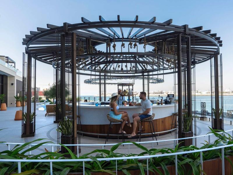 Dvě osoby sedí u kulatého baru pod otevřeným pavilonem s výhledem na moře venku.