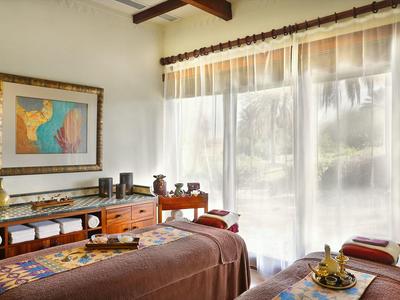 Bright massage room with two massage tables, wooden furniture, and large windows with curtains.