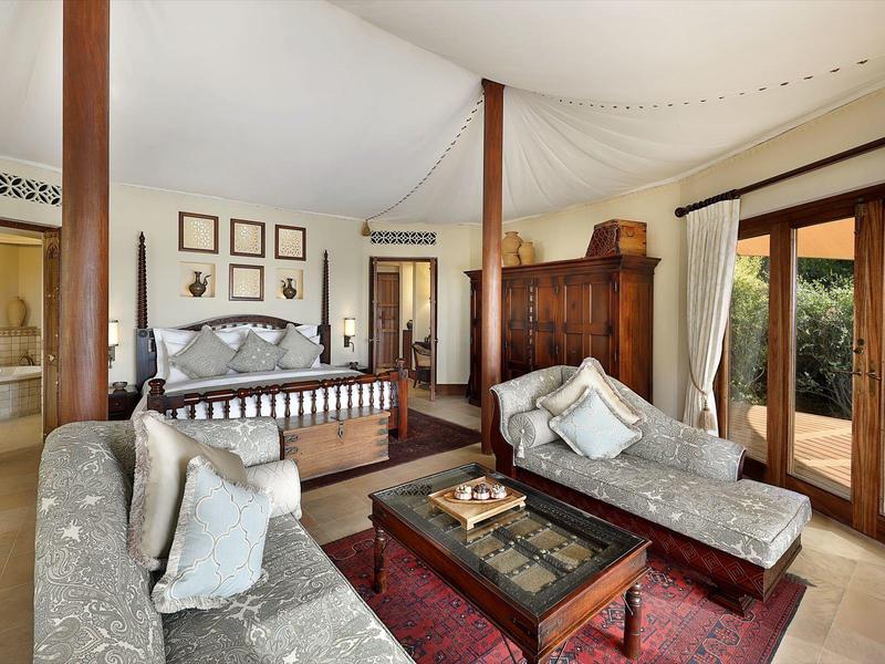 Elegant bedroom with four-poster bed, two lounges, and large windows overlooking greenery.