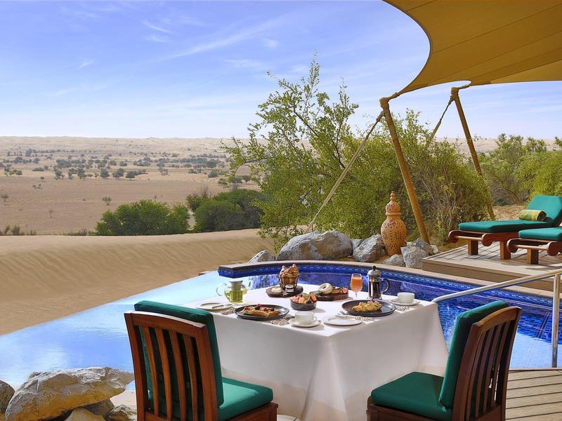 Terrace with set dining table and pool overlooking a sandy desert under a blue sky.