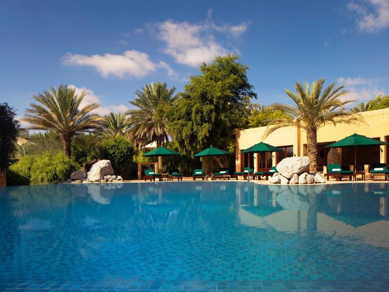 Clear blue pool with palm trees and sun umbrellas in front of hotel buildings.