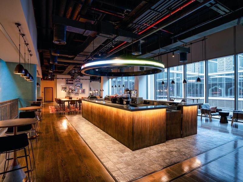 Modernes Café mit großem zentralem Tresen, Holzfußboden, hohen Fenstern und Stühlen aus Metall und Holz.