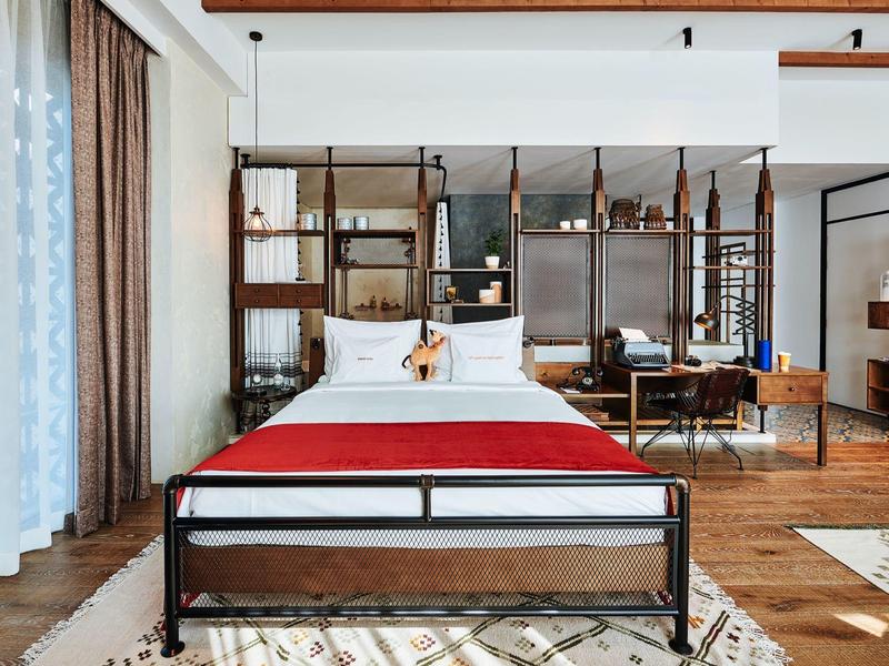 Modernes Schlafzimmer mit Eisenbett, roter Tagesdecke, Holzregal und großem Fenster mit Vorhängen.