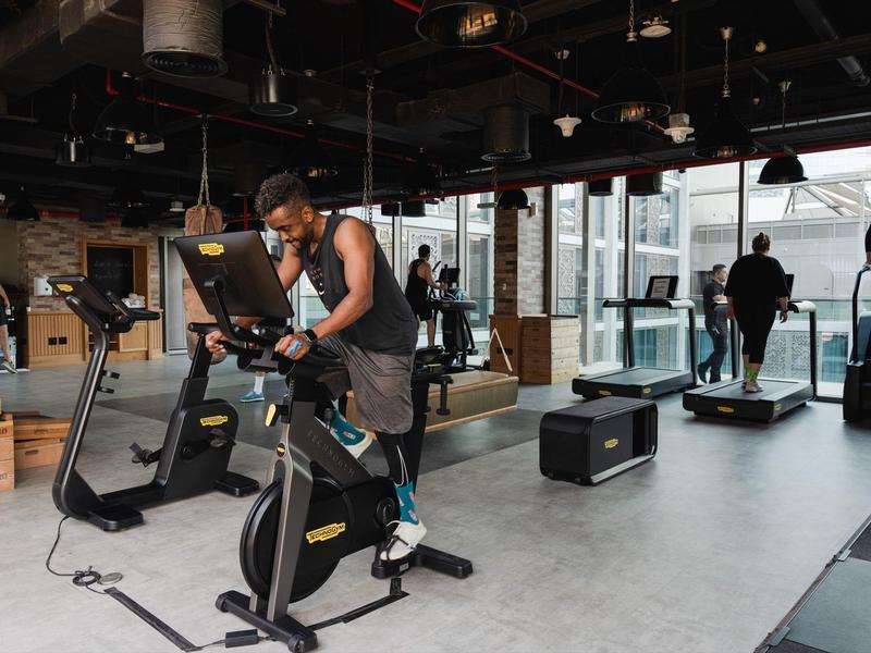 Moderne Fitnessstudio mit großen Fenstern, mehreren Trainierenden und verschiedenen Trainingsgeräten.