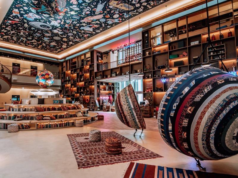 Großer, heller Raum mit bunten Teppichen, hängenden Körben, Holzregalen und verziertem Deckengewölbe.