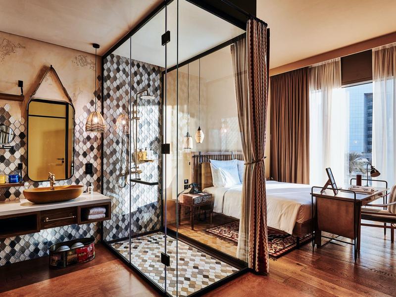 Modernes Hotelzimmer mit Glasdusche, Holzboden, floralem Fliesenmuster und großem Fenster.